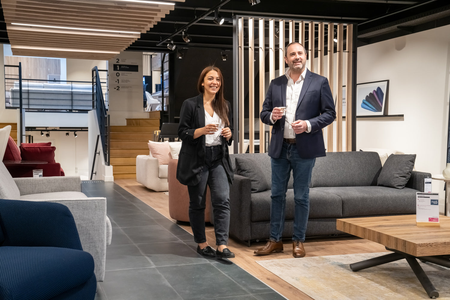 Deux personnes souriantes dans un showroom de meubles modernes, tenant des tasses, entourées de canapés et de tables en bois