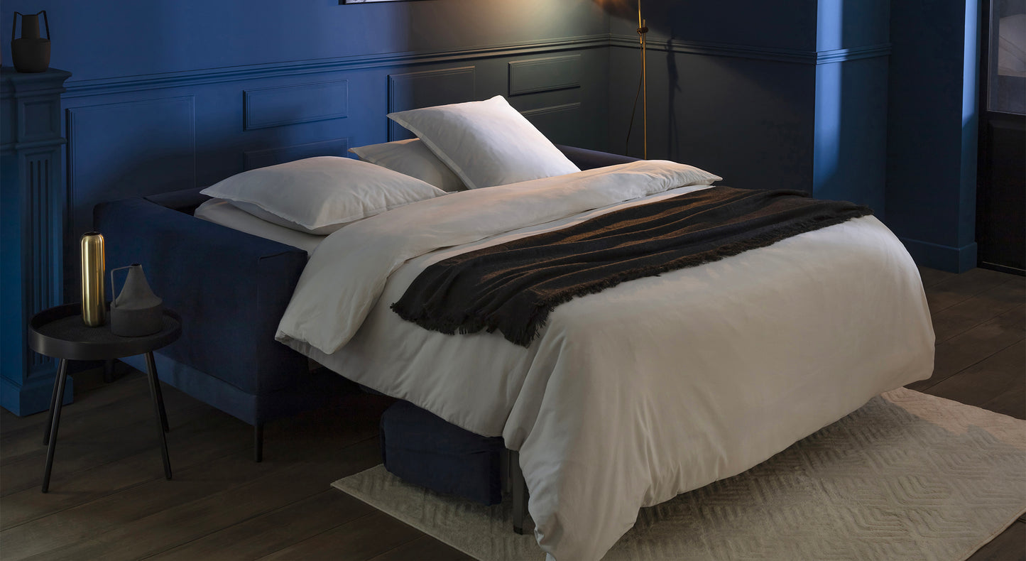 Canapé-lit en velours bleu océan avec couverture sombre, dans une pièce aux murs bleus et éclairage tamisé