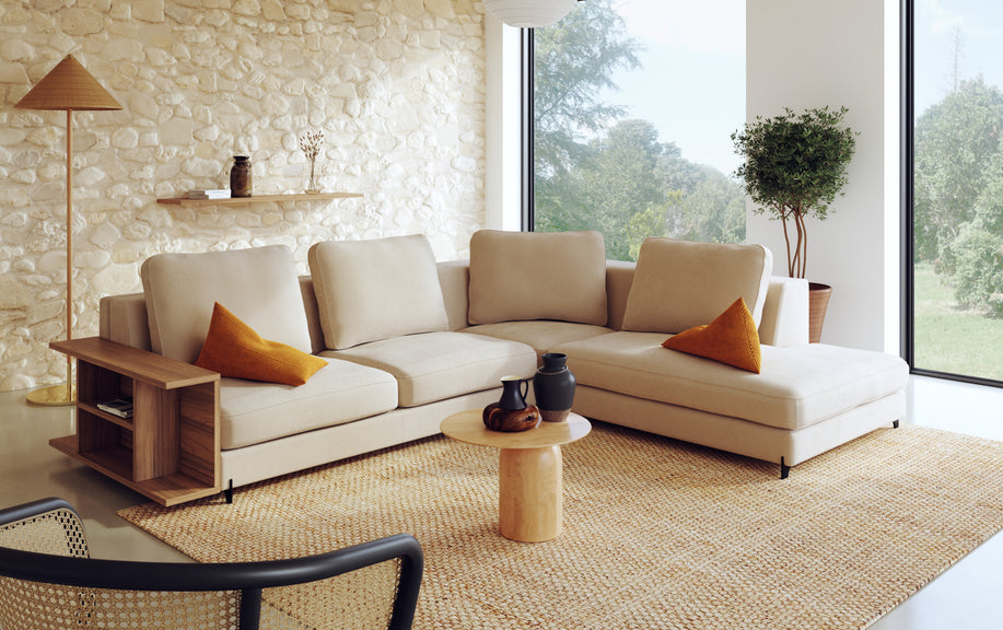 Canapé panoramique en tissu ivoire avec coussins orange, sur un tapis en jute, près d'une fenêtre et d'un mur en pierre. Ambiance chaleureuse