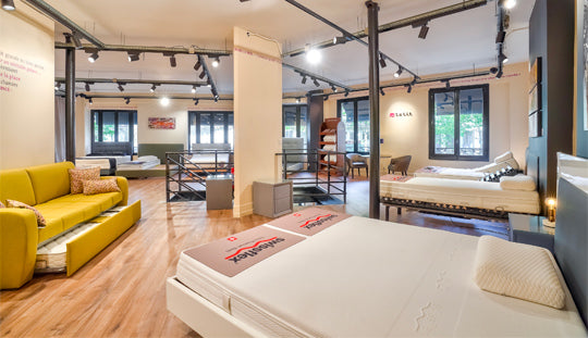 Salle d'exposition moderne avec plusieurs lits et un canapé, éclairage sur rails et grandes fenêtres