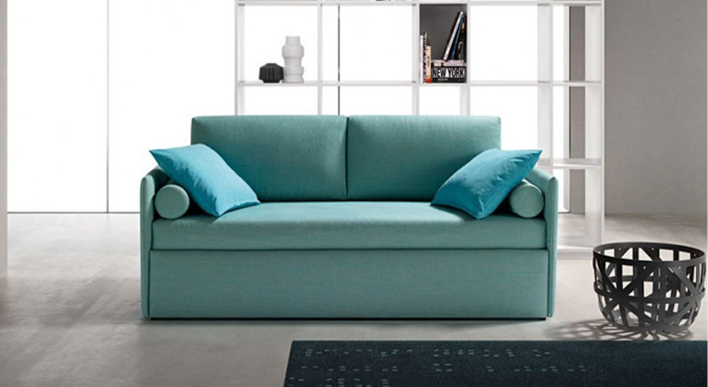 Sofa-lit gigogne en tissu bleu avec coussins, placé dans un salon moderne avec étagère blanche et panier décoratif