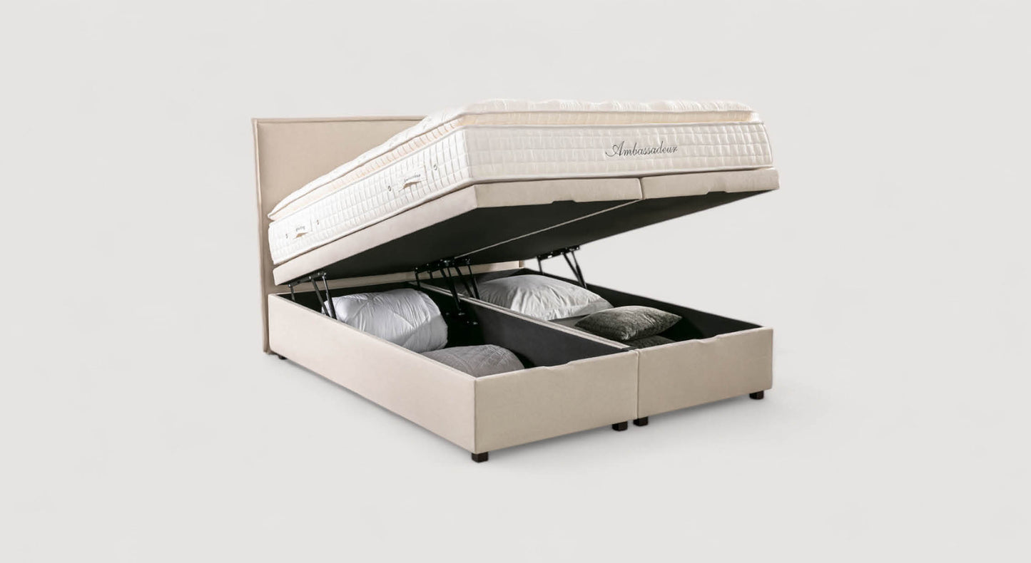 Lit coffre beige modèle Ouessant Luca avec matelas relevé, révélant un espace de rangement contenant des oreillers et des couvertures