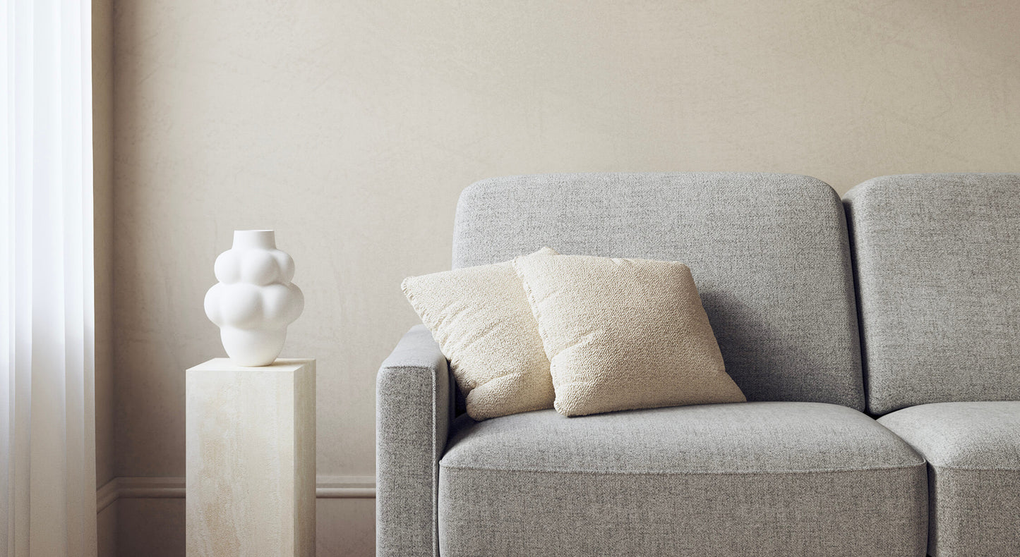 Canapé convertible gris en tissu Foster avec coussins beiges, à côté d'un vase blanc sculptural sur un socle, ambiance moderne et épurée