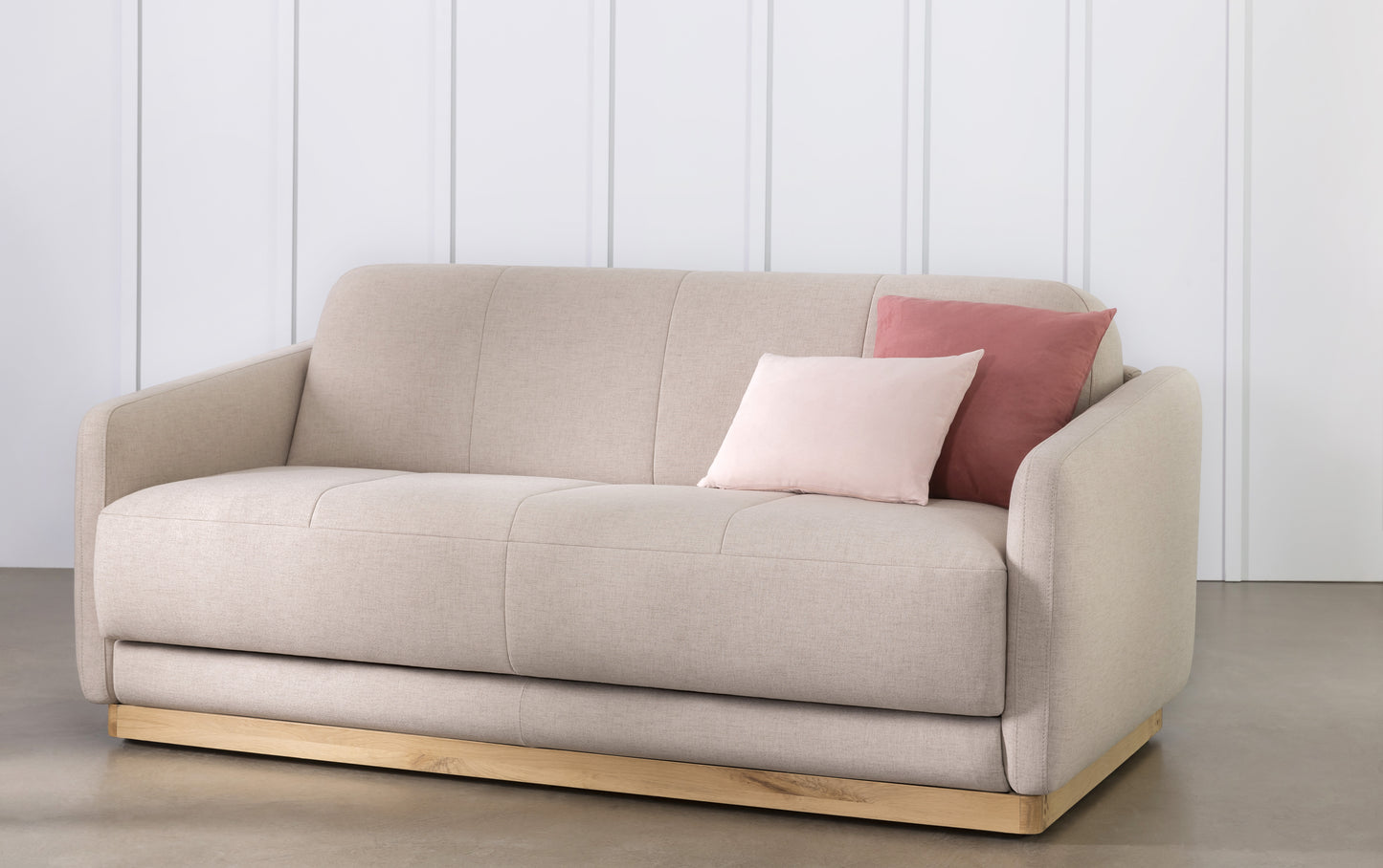 Canapé convertible en tissu mastic avec base en bois, deux coussins roses, style moderne et épuré