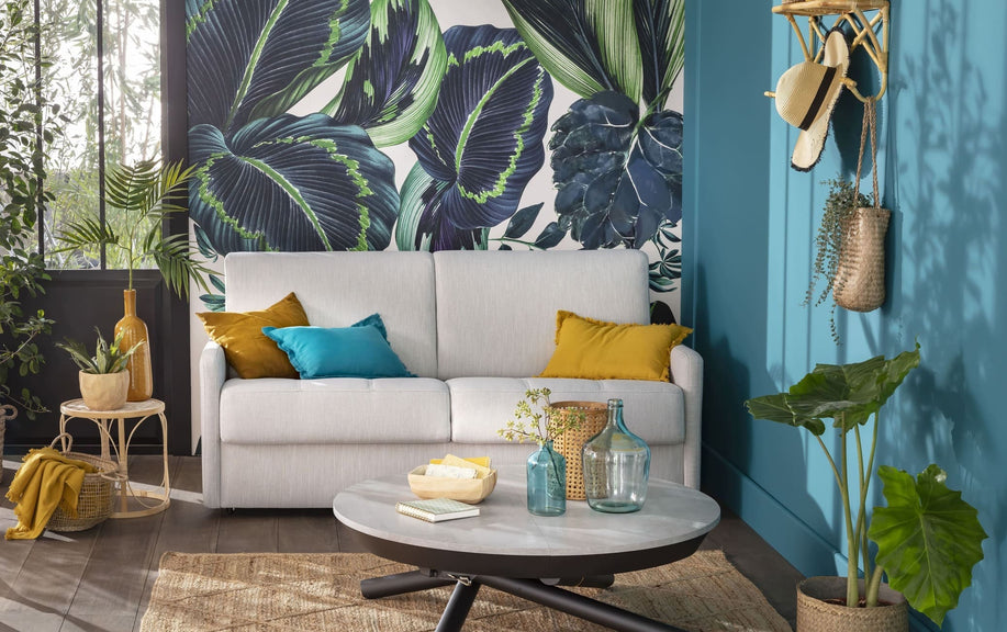 Canapé convertible Marlon en tissu chlorophile, entouré de coussins colorés, plantes et décor tropical, dans un salon lumineux et moderne