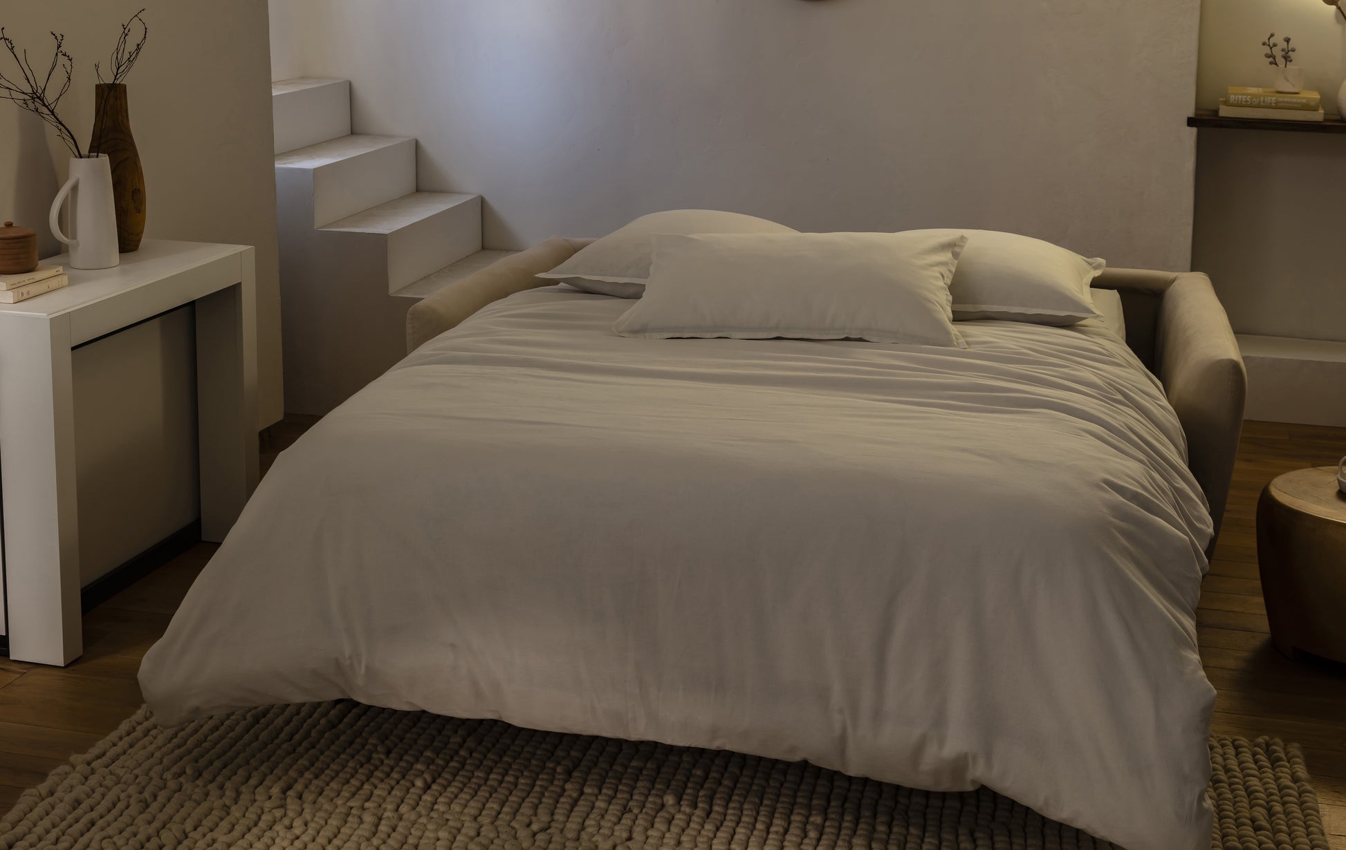 Canapé convertible beige en tissu, ambiance nuit, avec coussins, sur un tapis texturé dans un intérieur moderne et épuré