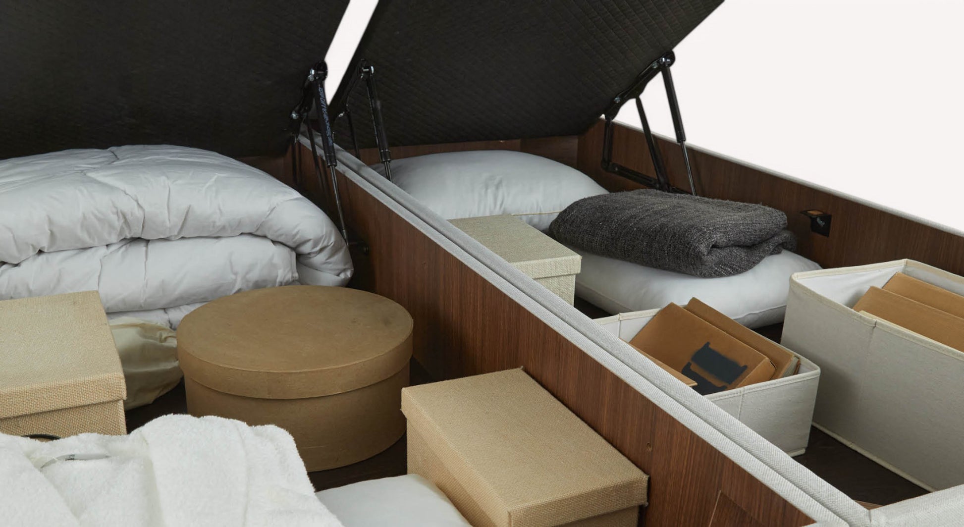 Espace de rangement sous un lit avec couettes blanches, boîtes de rangement en tissu et carton, et couverture grise pliée